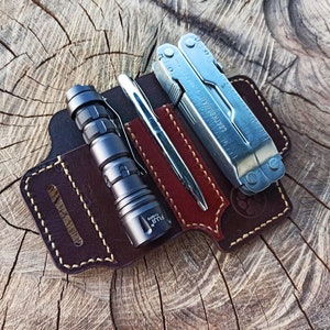 Puede incluir: Un soporte de herramientas multiusos de cuero marrón con una linterna, una herramienta multiusos y un bolígrafo. El soporte tiene un borde cosido y un bucle para el cinturón.