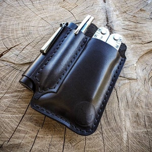 May include: A black leather multi-tool sheath with a pen and a small torch. The sheath is stitched and has a pocket for a multi-tool. The items are on a wooden surface.