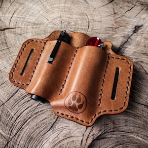 May include: A brown leather carrying case with two slots for holding a torch and a multi-tool. The case has a paw print design and is stitched with brown thread.