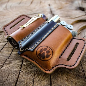 May include: A brown leather multi-tool sheath with a black accent and paw print design. The sheath holds a multi-tool and a flashlight. The leather is stitched with a contrasting thread and has a belt loop.
