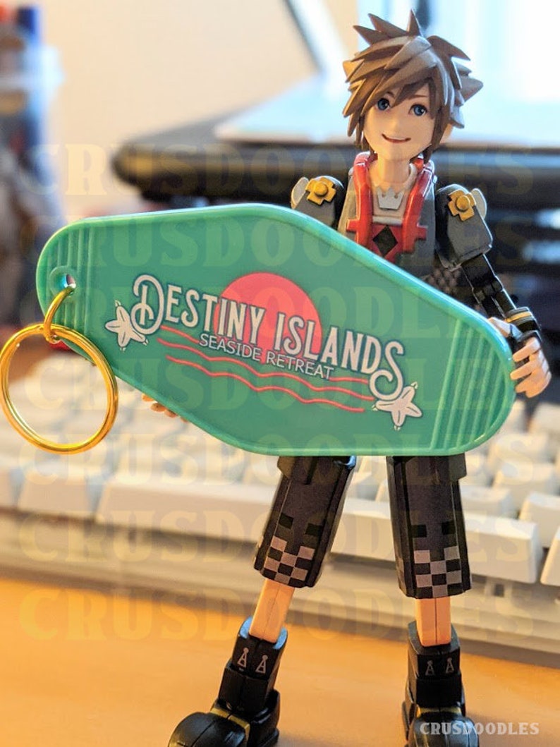 May include: A figurine of a character holding a teal motel-style keychain. The keychain reads "Destiny Islands Seaside Retreat" in white and red text. The character is wearing a black and gray outfit with a red vest.