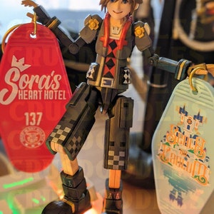 May include: An articulated action figure of a character from the Kingdom Hearts video game series, holding two keychains. One keychain is red with "Sora's Heart Hotel" printed on it, and the other is light blue with "Spooky Scream" printed on it. The figure is standing on a keyboard.