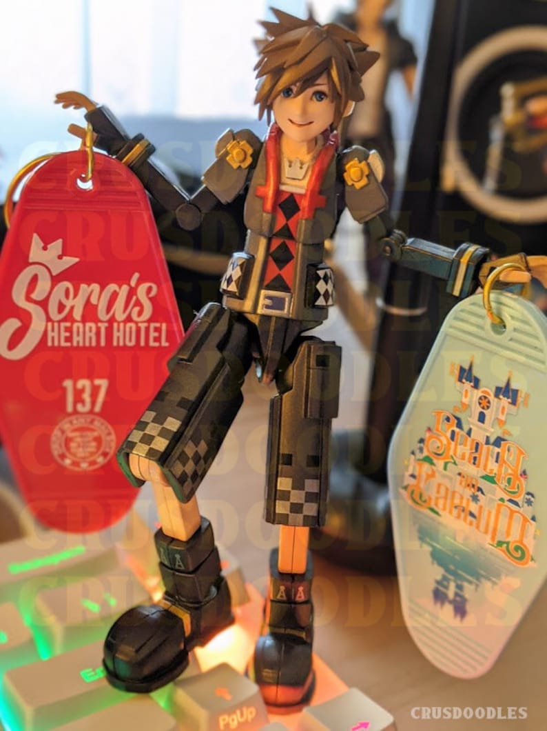 May include: An articulated action figure of a character from the Kingdom Hearts video game series, holding two keychains. One keychain is red with "Sora's Heart Hotel" printed on it, and the other is light blue with "Spooky Scream" printed on it. The figure is standing on a keyboard.