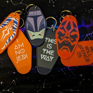 Puede incluir: Conjunto de seis llaveros naranja y negro con diferentes diseños de Star Wars. Los llaveros presentan imágenes de personajes y citas de las películas. Los llaveros tienen forma de llaves de motel.