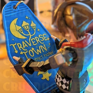 May include: A blue hotel keychain with gold lettering and a moon and lamp post design. The keychain reads "TRAVERSE TOWN" and "district hotel". A gold keyring is attached to the top. The keychain is held by a figurine.