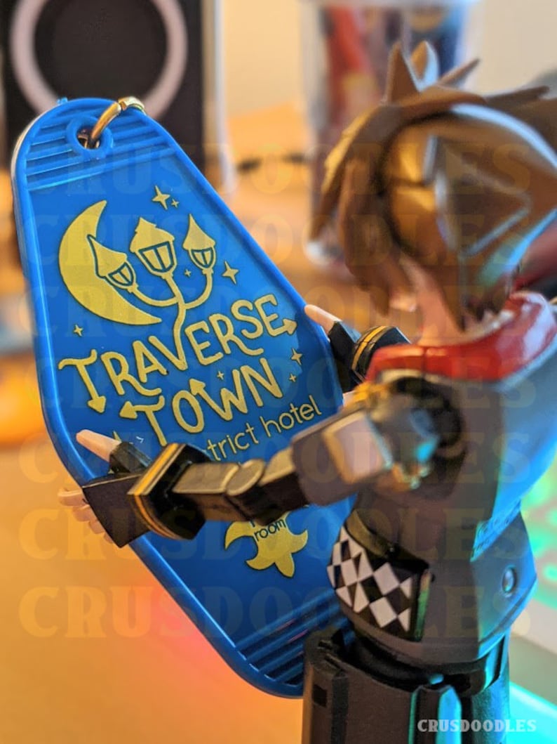 May include: A blue hotel keychain with gold lettering and a moon and lamp post design. The keychain reads "TRAVERSE TOWN" and "district hotel". A gold keyring is attached to the top. The keychain is held by a figurine.