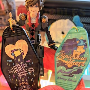 May include: A figurine of a character from the Kingdom Hearts video game series holding two keychains. One keychain is black with the text "The Hotel That Never Was" and the other is green with the text "The Mysterious Tower".