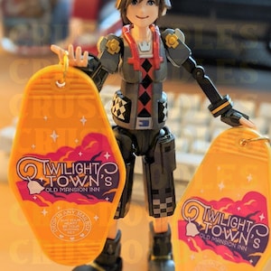 May include: A detailed action figure of a character holding two yellow keychains. The keychains have the text "Twilight Town's Old Mansion Inn" with a bell and cloud design. The figure is wearing a gray and black outfit.