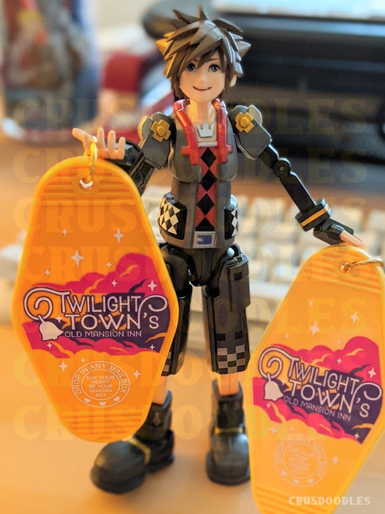 May include: A detailed action figure of a character holding two yellow keychains. The keychains have the text "Twilight Town's Old Mansion Inn" with a bell and cloud design. The figure is wearing a gray and black outfit.