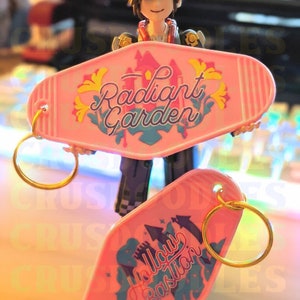 May include: Two pink motel-style keychains with gold keyrings. One keychain reads "Radiant Garden" with a castle graphic. The other keychain reads "Hollow Bastion" with a castle graphic. A figurine is holding the keychains.
