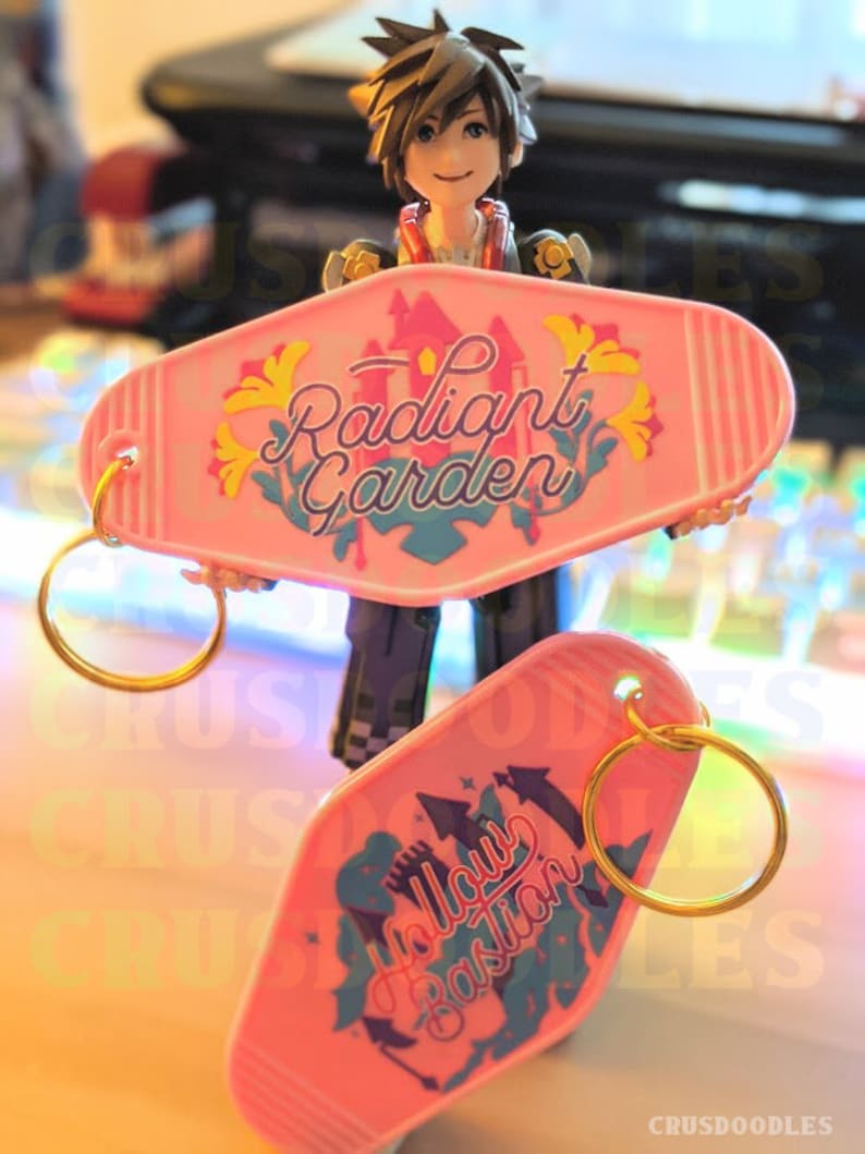 May include: Two pink motel-style keychains with gold keyrings. One keychain reads "Radiant Garden" with a castle graphic. The other keychain reads "Hollow Bastion" with a castle graphic. A figurine is holding the keychains.