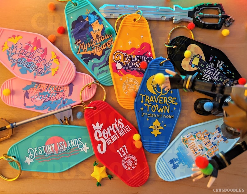 May include: A collection of colorful motel-style keychains with various designs and text. Keychains include "Radiant Garden", "Hollow Bastion", "The Mysterious Tower", "Twilight Town", "Destiny Islands", "Sora's Heart Hotel", and "Traverse Town".