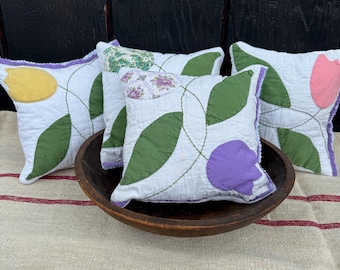 May include: Four square decorative pillows with white fabric, green leaves, and colorful flower accents in yellow, pink, and purple. The pillows are arranged in a wooden bowl on a linen surface.