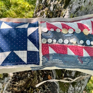 May include: A rectangular fabric pillow featuring a blue and white star quilt block on the left side and red and white striped fabric on the right. The pillow is embellished with rows of white and cream buttons and a strip of dark blue lace trim.