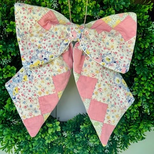 Op de afbeelding: Een decoratieve strik gemaakt van doorgestikte stof met een patchworkontwerp. Het patroon wisselt af tussen effen roze vierkanten en een crèmekleurige bloemenprint met kleine blauwe, gele en roze bloemetjes. Er is een dun touwtje aan bevestigd.