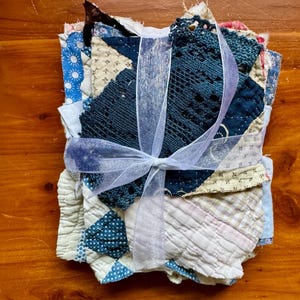 May include: A stack of assorted fabric scraps and textile pieces tied together with a white sheer ribbon. The bundle includes various patterns such as blue and white sun motifs, cream-colored geometric prints, and a dark blue crocheted lace piece.