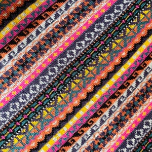 K&ouml;nnte beinhalten: Nahaufnahme eines farbenfrohen Textils mit diagonalem Streifenmuster. Das Design zeigt B&auml;nder in Pink, Gelb, Orange, T&uuml;rkis und Lila mit geometrischen und tribal inspirierten Motiven. Der Stoff hat ein strukturiertes Aussehen.
