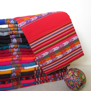 May include: A stack of colorful woven textiles, featuring a vibrant red textile on top with intricate geometric patterns in various colors. The textiles are folded and stacked, showcasing a variety of hues. A decorative sphere with floral designs sits nearby.