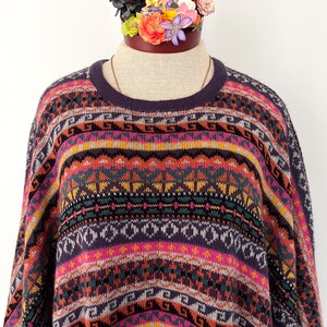 K&ouml;nnte beinhalten: Ein farbenfroher Pullover mit Muster und Rundhalsausschnitt. Der Pullover hat geometrische und gestreifte Designs in Lila, Rosa, Orange und T&uuml;rkis. Das Kleidungsst&uuml;ck wird auf einer Schaufensterpuppe mit einem Blumenarrangement oben pr&auml;sentiert.