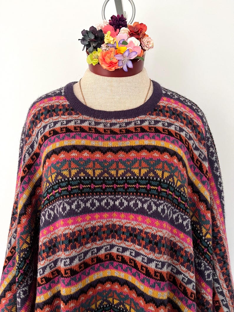 K&ouml;nnte beinhalten: Ein farbenfroher Pullover mit Muster und Rundhalsausschnitt. Der Pullover hat geometrische und gestreifte Designs in Lila, Rosa, Orange und T&uuml;rkis. Das Kleidungsst&uuml;ck wird auf einer Schaufensterpuppe mit einem Blumenarrangement oben pr&auml;sentiert.