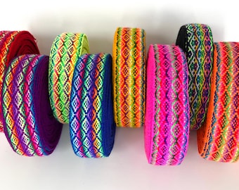 May include: Eight rolls of woven fabric trim arranged in a row. Each roll features a vibrant, multi-colored geometric diamond pattern. The colors include bright pink, orange, yellow, lime green, purple, blue, and red, with intricate detailing.