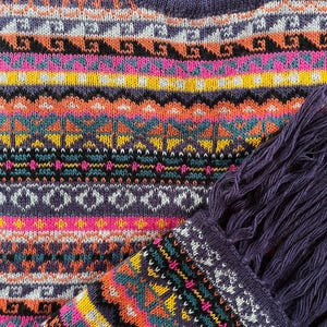 K&ouml;nnte beinhalten: Nahaufnahme eines farbenfrohen Strickpullovers mit geometrischem Muster. Der Pullover hat einen V-Ausschnitt und einen Fransen-Schal. Das Design umfasst Farbt&ouml;ne von Lila, Rosa, Orange, Gelb und T&uuml;rkis. Das Strickwaren ist wahrscheinlich handgefertigt.
