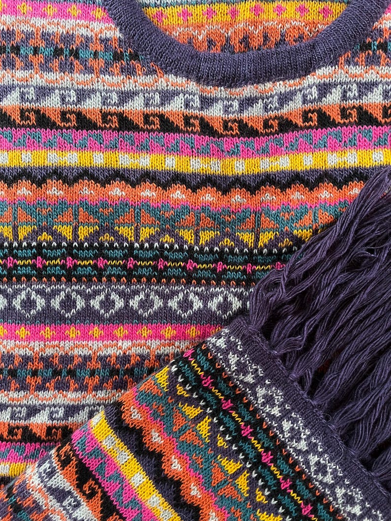 K&ouml;nnte beinhalten: Nahaufnahme eines farbenfrohen Strickpullovers mit geometrischem Muster. Der Pullover hat einen V-Ausschnitt und einen Fransen-Schal. Das Design umfasst Farbt&ouml;ne von Lila, Rosa, Orange, Gelb und T&uuml;rkis. Das Strickwaren ist wahrscheinlich handgefertigt.