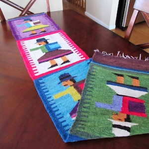 May include: A colourful table runner with a repeating pattern of figures in traditional clothing. The runner is made of woven fabric and features shades of purple, pink, blue, green, and white.