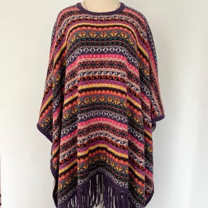 K&ouml;nnte beinhalten: Ein farbenfrohes Poncho mit geometrischem Muster in Lila, Rosa, Gelb und Schwarz. Der Poncho hat einen runden Ausschnitt und Fransen am unteren Rand. Das Kleidungsst&uuml;ck wird auf einer Schaufensterpuppe pr&auml;sentiert.