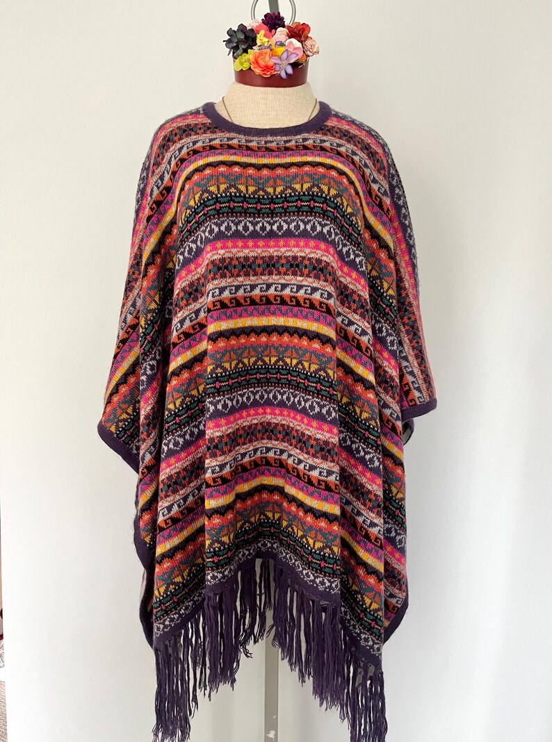 K&ouml;nnte beinhalten: Ein farbenfrohes Poncho mit geometrischem Muster in Lila, Rosa, Gelb und Schwarz. Der Poncho hat einen runden Ausschnitt und Fransen am unteren Rand. Das Kleidungsst&uuml;ck wird auf einer Schaufensterpuppe pr&auml;sentiert.