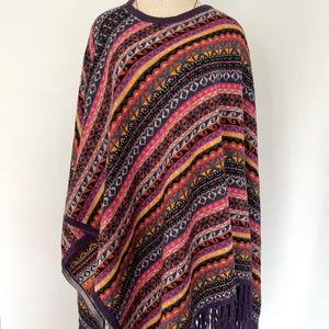 K&ouml;nnte beinhalten: Ein farbenfrohes Poncho mit einem geometrischen Muster in Lila, Rosa, Gelb und Schwarz. Der Poncho hat einen Fransenbesatz und wird auf einer Schaufensterpuppe pr&auml;sentiert. Das Design zeigt Streifen und Rauten.