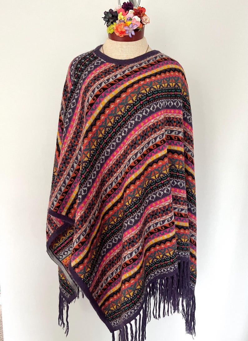 K&ouml;nnte beinhalten: Ein farbenfrohes Poncho mit einem geometrischen Muster in Lila, Rosa, Gelb und Schwarz. Der Poncho hat einen Fransenbesatz und wird auf einer Schaufensterpuppe pr&auml;sentiert. Das Design zeigt Streifen und Rauten.