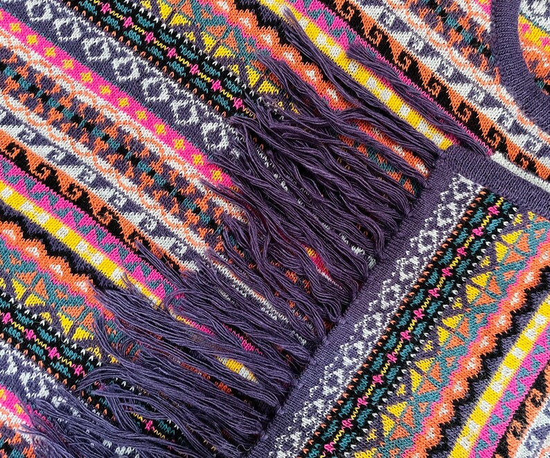 K&ouml;nnte beinhalten: Nahaufnahme eines farbenfrohen Strickschals mit geometrischem Muster. Der Schal weist eine lebendige Mischung aus Farben auf, darunter Lila, Rosa, Orange, Gelb und Wei&szlig;. Der Schal hat einen Fransen aus lila Garn.
