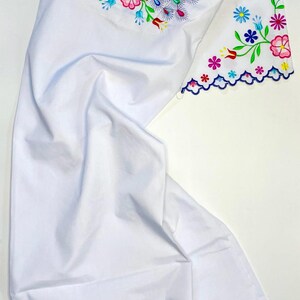 May include: White fabric with colorful embroidered peacock and floral designs. The embroidery features blue peacocks, pink, yellow, and blue flowers, and green leaves. The edges are scalloped and trimmed with a blue border.