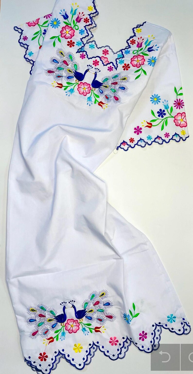 May include: White fabric with colorful embroidered peacock and floral designs. The embroidery features blue peacocks, pink, yellow, and blue flowers, and green leaves. The edges are scalloped and trimmed with a blue border.