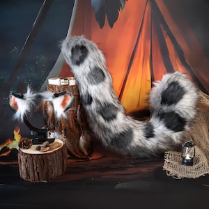 Può includere: Un set cosplay in finta pelliccia con una coda maculata, una fascia per le orecchie da gatto e un collare bianco. La coda è grigia e nera, e le orecchie sono bianche, grigie e arancioni. Il set è esposto su una superficie di legno con uno sfondo rustico.