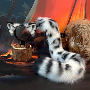 May include: A set of faux fur animal ears on a black headband and a long, matching faux fur tail. Both pieces feature a white base with black spots and stripes, mimicking a snow leopard pattern. The ears are upright and the tail is curved.