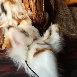 Op de afbeelding: Een paar pluizige, imitatiebont kattenoren in wit, bruin en beige. De oren hebben een roze binnenkant en zijn bevestigd aan een zwarte draad. De achtergrond bevat een houten element met veren en kralen.