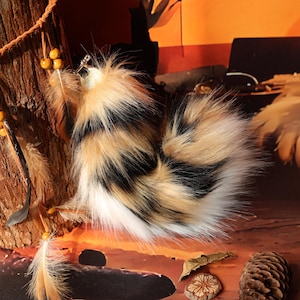 May include: A fluffy, faux fur tail in a mix of black, tan, and white. The tail is round and has a clip attached. The image also shows decorative feathers and wooden beads.