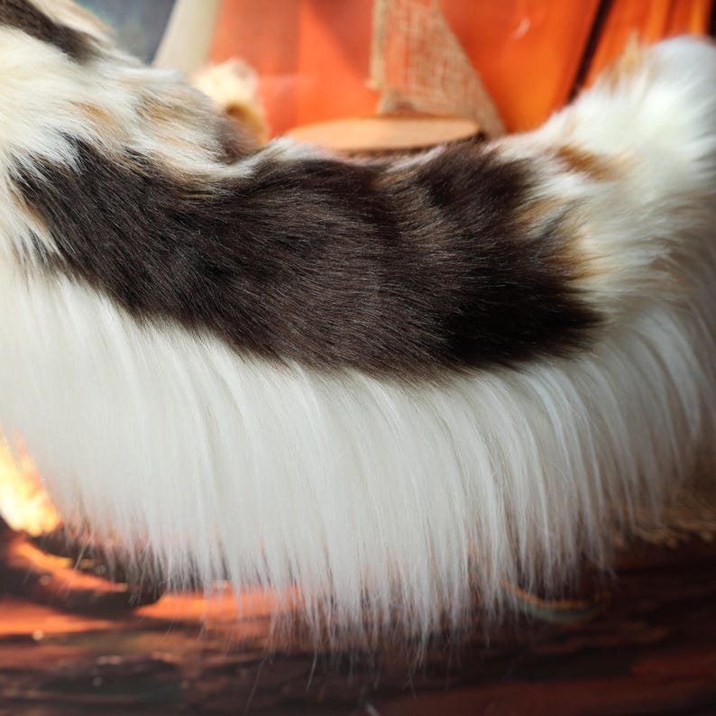 Op de afbeelding: Close-up van een pluizig, imitatiebont item met een kleurenpatroon van wit, bruin en bruin. Het bont is lang en zacht, met een mix van kleuren die een textuur cre&euml;ert. Het item lijkt een staart of een vergelijkbaar accessoire te zijn.