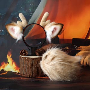 Peut inclure: Un serre-tête avec des oreilles d'animal en fausse fourrure beige et de petits bois, accompagné d'une queue en fausse fourrure beige assortie. Le serre-tête est présenté sur un support en bois cylindrique et la queue est posée devant.
