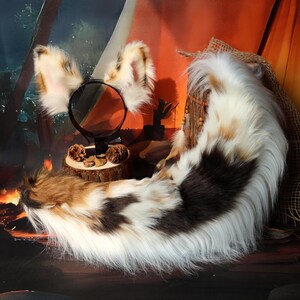 Op de afbeelding: Een set van nepbont kattenoren op een zwarte hoofdband en een bijpassende staart. De oren en de staart hebben een mix van witte, bruine en bruine vacht. De staart is lang en pluizig.