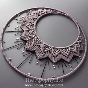 May include: A macrame wall hanging in the shape of a crescent moon with a geometric pattern. The moon is made of light purple cord and is decorated with small, clear crystals.