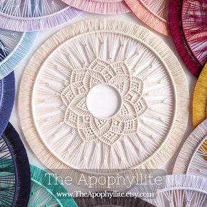 May include: A collection of macrame wall hangings in various colors, including white, blue, red, and yellow. The hangings are circular and feature intricate knot patterns. The text "The Apophyllite www.TheApophyllite.etsy.com" is visible at the bottom of the image.