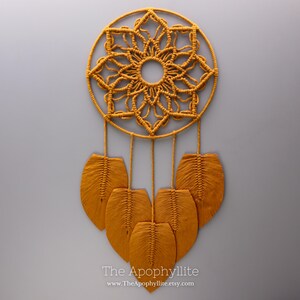 May include: Mustard yellow macrame dream catcher with a circular design and leaf-shaped accents. The dream catcher features a central floral pattern and hanging leaves. The text "The Apophyllite" and website address are visible at the bottom.