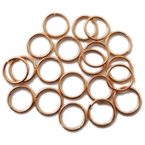 Pode incluir: Uma coleção de pequenos anéis de metal cor de ouro rosa. Os anéis são circulares e parecem sem costura, com uma ligeira abertura visível em alguns. As peças de joalharia estão espalhadas sobre um fundo branco, sugerindo um tema de artesanato ou joalharia.