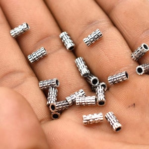 May include: A close-up of a hand holding a pile of silver metal spacer beads. The beads are cylindrical and have a textured surface.