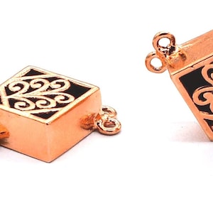 May include: A rose gold tone metal clasp with a square design featuring a black filigree pattern. The clasp has two loops on each side for attaching to a necklace or bracelet.