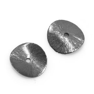 Peut inclure: Deux perles en forme de disque en métal gris foncé, texturées, avec un trou central. Les perles présentent un motif radial émanant du centre. Les perles sont posées sur un fond blanc.