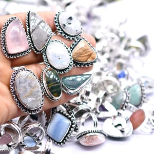 May include: A collection of gemstone rings with silver-colored settings. The rings feature various stone shapes and colors, including pink, white, gray, and green. The rings are displayed against a white background.